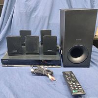 Sony 5.1 channel surround sound system components including amplifier/DVD unit, 5 satellite speakers, subwoofer, remote control, and cables on a blue cloth surface.