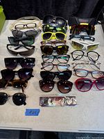 Top down view of 16 pairs of sunglasses and eyeglasses arranged on a white surface, some with cases included. Various frame colors and lens types visible.