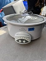 White Rival Crock Pot slow cooker with transparent glass lid placed outdoors on a surface, control panel visible and operational.