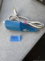 Photo showing blue electric engraving pen with power cord attached and two-prong plug, resting on a grey surface with #12 label nearby.