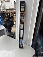 Photo of a black 24-inch magnetic top-read level with yellow liquid vials in a vertical position, leaning against a white doorframe outdoors with assorted background items.