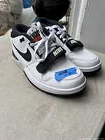 Photo shows pair of white and black Nike running shoes with 'NIKE' strap across laces and size note on blue tape on toe box.