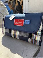 Front view of folded picnic blanket in plaid pattern with navy blue flap and red label with text 'Great Canadian Outdoors Blanket'.