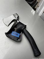 Single small axe with metal blade and black rubber handle, protected by a black leather sheath with white stitching and snap closure.