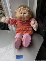 Front view of the vintage Cabbage Patch Kids doll sitting, showing face, hair, and original orange jumper dress with pink and orange striped shirt and pink pants with shoes.