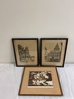 Two framed city landscape art prints leaning against a wall with one flat laying in front on a white surface.