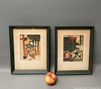 Two framed Asian art lithographs side by side with an apple for scale. Both frames are dark green with cream mats.
