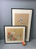 Two framed Asian bird paintings on silk with flower motifs, black bamboo-style frames, set against grey wall with an apple for scale.