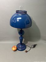 Full view of the blue metal Art Deco table lamp with dome shade and glass chimney, alongside an apple for scale.
