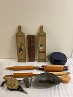 Various Country Kitchen items arranged on a white table: two painted wooden breadboards, carved wooden butter mold, three wooden rolling pins, dark blue cast iron pot with lid, brass hand bellows, metal funnel, metal dough cutter, small wooden container.