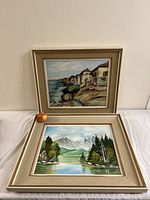 Both landscape oil paintings, one resting upright showing a coastal village scene and the other lying down showing a mountain lake scene.