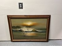 Framed oil painting of seascape with crashing waves resting against a plain white wall, showing full view of painting and frame.