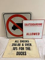 Three signs placed together: No Trucks prohibition sign, Skateboarding Allowed sign, and Ducks check notice sign.