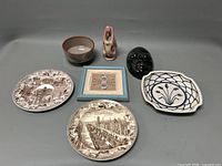 Overview of all lot items including two transferware plates, small bowl, two pottery figurines, framed textile piece, and ceramic dish