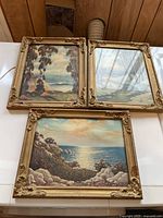 Three framed small prints with coastal landscape scenes laid on a table, showing ornate gold frames.