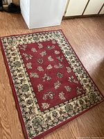 Photo showing the full rectangular area rug on wooden floor, highlighting red central field and beige floral border.