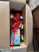 Top-down view showing a box filled with various cleaning supplies including a red feather duster, spray bottles, and boxed household items.