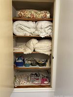 Photo of closet shelves showing neatly folded floral, white and cream colored blankets and throws on the top three shelves. Below these are multiple baskets containing various small household items including cords, light bulbs, and miscellaneous tools. Bottom shelf has packaged sheet grippers, light bulbs, and hot/cold therapy packs.