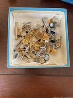 Assorted cufflinks and tie clips in a box showing various gold-tone and silver-tone pieces with different designs and some with colored stones.