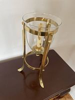 Close-up photo of the decorative candle display holder showing a glass hurricane supported by a gold-toned metal stand with a white candle inside.