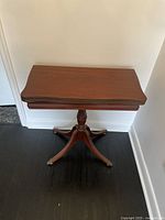 Frontal view of wooden folding table with curved edged top folded down, pedestal base and four curved legs with metal capped feet.