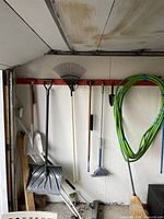 Photo of various garden tools hanging on a wall including rakes, shovels, broom, and a coiled garden hose