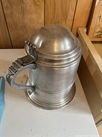 Metal beer stein with curved handle and domed, hinged lid shown from side angle