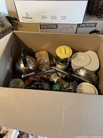 Opened box containing kettle, strainers, handheld graters, pepper grinder, plastic containers with lids, and miscellaneous kitchen utensils.