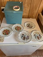 Five ceramic lace edge dessert plates in a set; three with peacock motifs and two with floral designs, pictured with a blue box.