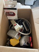 Top-down view of mixed electronics lot inside an open cardboard box showing GNC AccuTrack box, power strip, clock radio, flashlight, and other items.