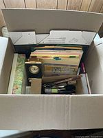 Top view of a boxed lot showing an assortment of office materials including a heavy clock, folders, papers, and various assorted items.