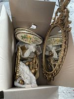 Box containing ornate framed oval mirror, floral ceramic plate, and gold-toned decorative items