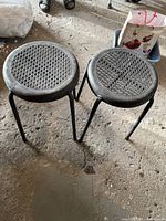 Two black metal frame outdoor stools with circular woven seats shown on a rough concrete floor.