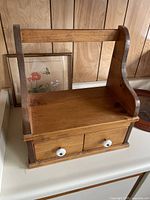 Small pine wood shelf with two front drawers and white knobs, showing front and side views against a wood panel wall and a countertop.