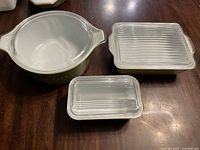 Three Pyrex green and clear casserole dishes with clear ribbed lids on a wooden surface