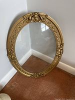 Full view of the oval vintage picture frame with gold ornate design and rounded glass, standing on a carpeted floor in a corner.