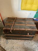 Closed view of the steamer trunk showing the exterior wood slats and leather strap over the top, with aged and worn surface.
