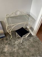 Full view of white wrought iron makeup table with glass top and matching chair with black cushion on carpeted floor against off-white wall.