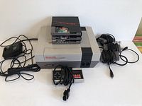 Front view of NES console with Tecmo Bowl game cartridge inserted, two additional cartridges stacked on top, controller, power adapter, and cables laid out around.
