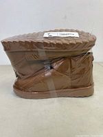 Side profile of stacked pair of brown winter ankle boots wrapped in plastic showing flat sole and synthetic upper.