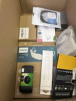 Top view of box showing multiple electronics accessories including Philips headphones box, disposable camera, phone cases, charger cable, and screen protector.