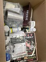 Box with assorted items including a Keurig accessory box, cylindrical metal part, and small bottles with miscellaneous items below.
