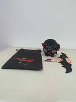 Menace Mauler Longpire plushie, black branded drawstring bag, bat-shaped wings and guitar-shaped accessory laid flat on surface.