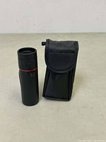 Black monocular with red ring standing beside its black carrying case with flap.