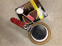 Close-up of orbital sander with red handle beside unopened Tusk brand sandpaper pack.
