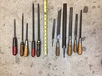 Top-down view of 12 metal hand files with various handles laid next to measuring tape for size reference on concrete floor.