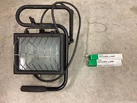 Full view of the halogen work light with handle and protective grill, alongside two extra halogen lamp bulbs in green and white boxes.