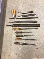 Top-down view of 10 metal hand files with wood handles arranged on a beige floor, showing various sizes and shapes.