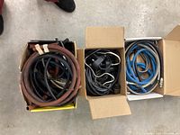Three boxes each holding coiled hoses of various sizes, colors and types including rubber and plastic materials with some attached fittings, photographed from above on a concrete floor.