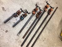 Set of six metal pipe clamps laid on a concrete floor. Clamps have orange and blue clamp heads, varying lengths and visible rust and wear marks.